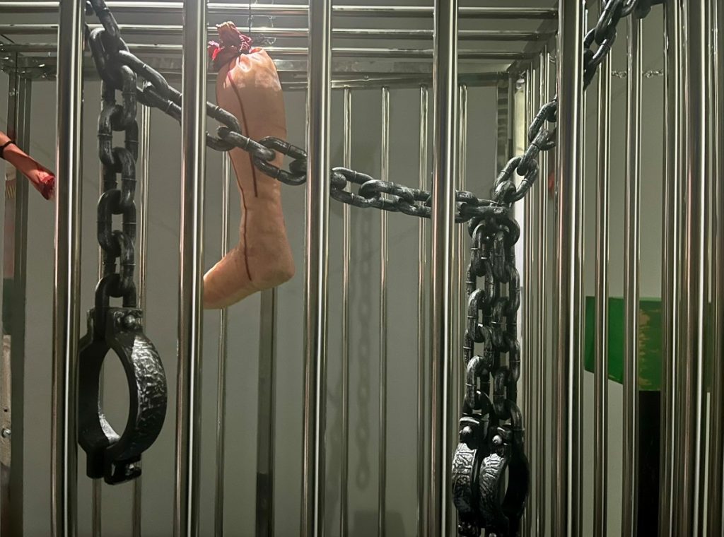 A metal cage with chains and handcuffs attached to the bars. A realistic-looking fake leg, tied with red string, hangs inside the cage. The lighting is dim and the atmosphere appears eerie.