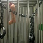 A metal cage with chains and handcuffs attached to the bars. A realistic-looking fake leg, tied with red string, hangs inside the cage. The lighting is dim and the atmosphere appears eerie.