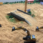 A miniature construction site diorama with toy excavators, a partially built bridge, a crane, caution signs, and a sandy, rocky terrain.