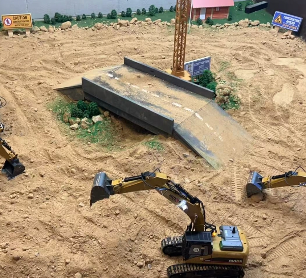 A miniature construction site diorama with toy excavators, a partially built bridge, a crane, caution signs, and a sandy, rocky terrain.