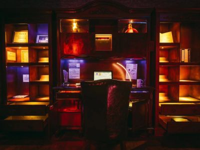 A dimly lit, vintage wooden desk and shelves with warm accent lights. An armchair faces a laptop on the desk, surrounded by documents, a mug, open drawers, and decorative items on the shelves.