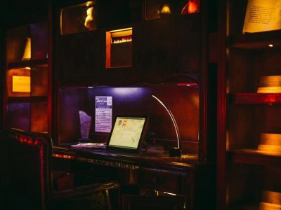 A dimly lit wooden desk with a computer monitor, modern desk lamp, papers, and an armchair, surrounded by shelves with books and warm ambient lighting.