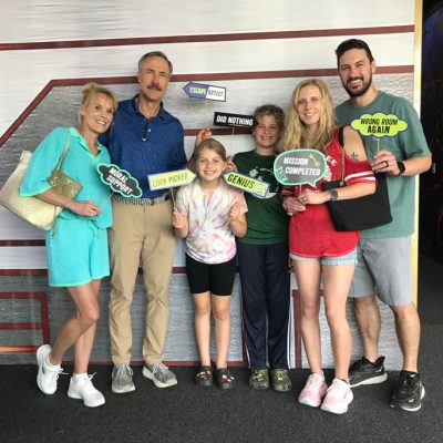 A group of six people, including three adults and three children, smile and hold colorful escape room signs with phrases like Mission Completed and Genius while posing together indoors.