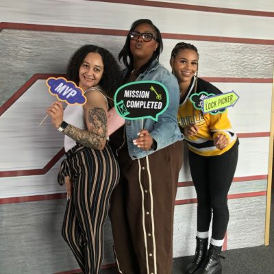 Three women stand smiling and posing indoors, each holding colorful signs that read MVP, Mission Completed, and Master Lock Picker against a modern, striped wall background.