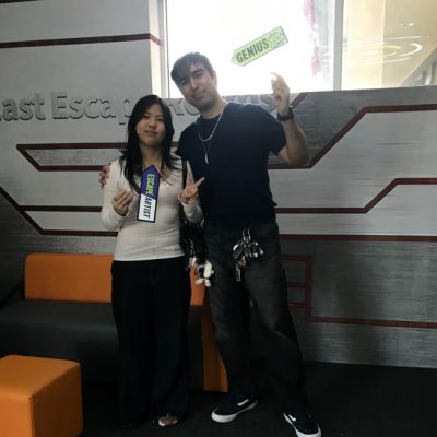 Two people stand indoors, holding signs that say GENIUS and GENIUS TEAM. They are smiling and making peace signs, with a wall that reads Escape partially visible behind them.