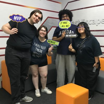 Four people stand together smiling in a modern room with orange and gray seating, holding colorful signs that say MVP, It was all me!, Genius, and I was told there would be no math.