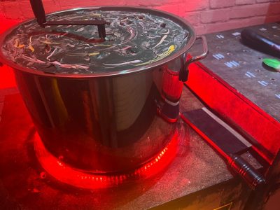 A large metal pot with a lid sits on a surface illuminated by red lights. The pot reflects nearby objects, and a flashlight and small tools are beside it. A brick wall and control panel are in the background.