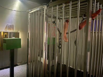 A dimly lit room with a large metal cage containing chained, realistic-looking fake severed arms and legs hanging from the bars. There are green and black boxes and chains attached to the cage.