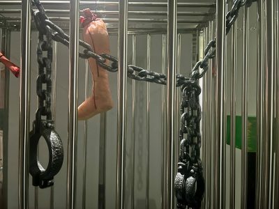 A metal cage with chains and handcuffs attached to the bars. A realistic-looking fake leg, tied with red string, hangs inside the cage. The lighting is dim and the atmosphere appears eerie.