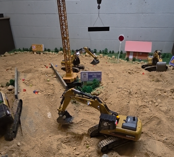 A miniature construction site model with toy excavators, a crane, sand, small signs, and a pink building, set against a gray wall.