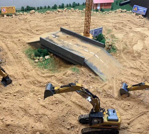 A miniature construction site diorama with toy excavators, a partially built bridge, a crane, caution signs, and a sandy, rocky terrain.